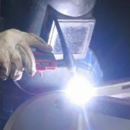 Spawanie metodą TIG: spawacz w masce ochronnej łączy metalowe elementy, widoczne iskry i błysk spawania, w tle fragment niebieskiej koszuli w kratę.