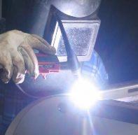 Spawanie metodą TIG: spawacz w masce ochronnej łączy metalowe elementy, widoczne iskry i błysk spawania, w tle fragment niebieskiej koszuli w kratę.