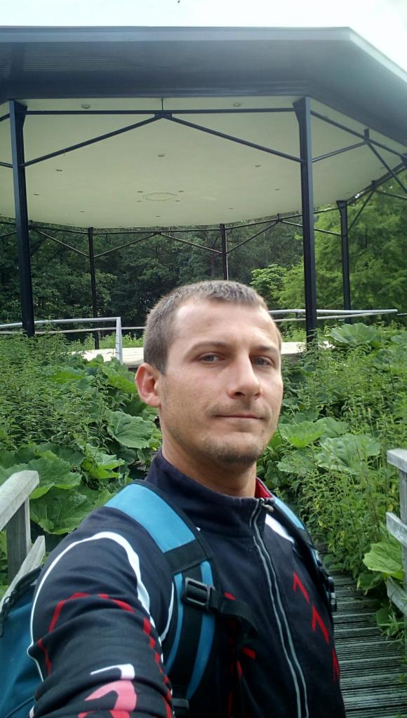 Mężczyzna z plecakiem na tle altany w parku, otoczonej bujną zielenią. Ujęcie z dołu, widoczny fragment drewnianego mostku.