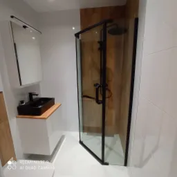 Nowoczesna łazienka z kabiną prysznicową z czarnymi profilami, umywalką nablatową w czarnym kolorze na drewnianym blacie i białymi płytkami na ścianach.