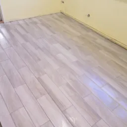 Ułożona podłoga z płytek imitujących drewno w jasnym odcieniu szarości, widoczne fragmenty żółtej ściany z wystającymi kablami.