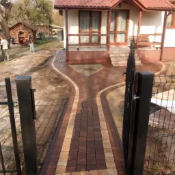 Nowo ułożony chodnik z kostki brukowej w dwóch odcieniach brązu, rozwidlający się w kształcie litery Y przed wejściem do domu z czerwonym dachem, widok zza otwartej, czarnej furtki.