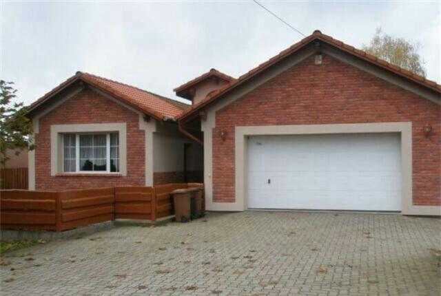 Dom jednorodzinny z garażem w Piasku, z czerwoną ceglaną elewacją i białymi obramowaniami okien i drzwi. Widoczne kosze na śmieci przy drewnianym płocie.
