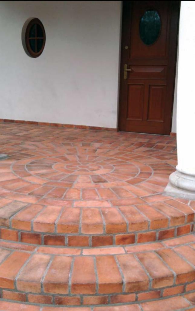 Wejście do budynku z wyłożonym cegłą schodkowym podejściem i okrągłym oknem na białej ścianie, ciemne drzwi wejściowe z owalną szybą.