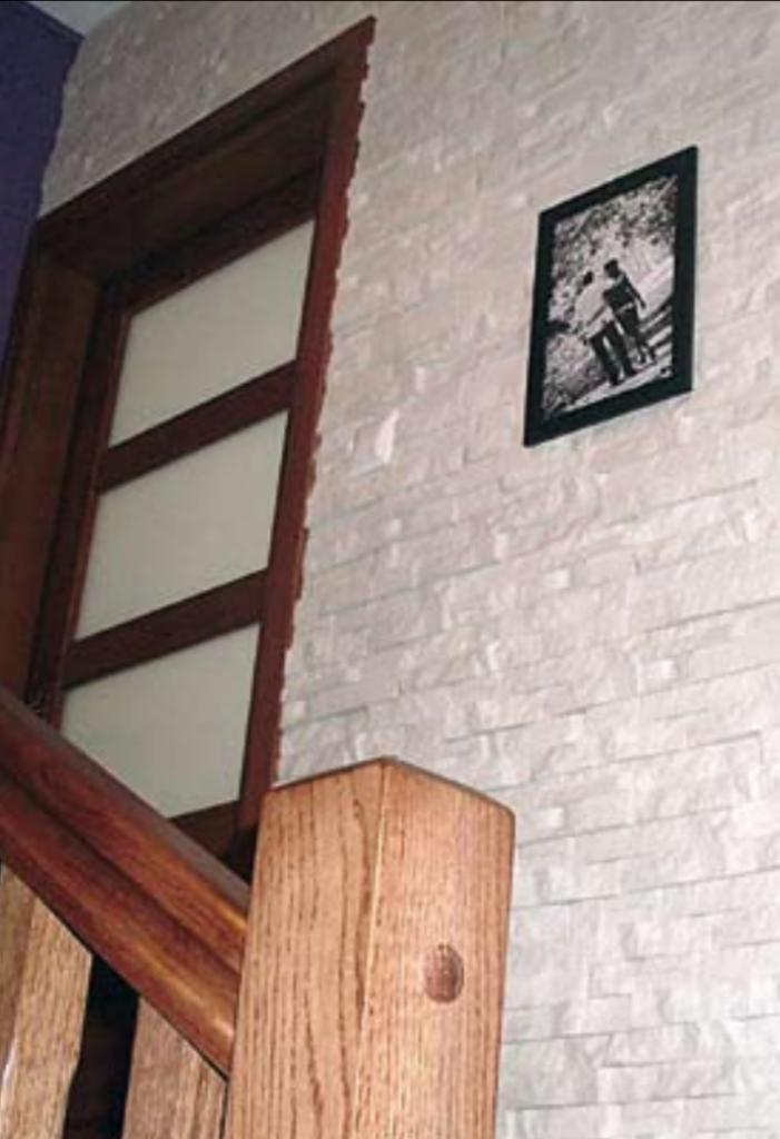 Ściana wewnętrzna wykończona dekoracyjnym białym kamieniem, z widoczną drewnianą ramą drzwi z przeszkleniami oraz czarno-białym zdjęciem w ramce, fragment drewnianej balustrady schodów w dolnym...