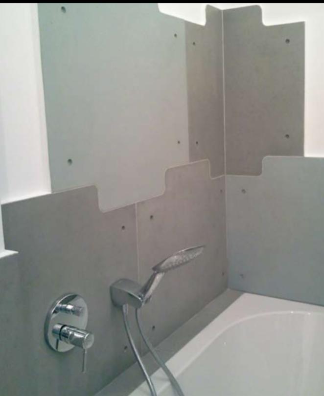 Montaż szarych paneli ściennych z otworami, imitujących beton, nad białą wanną z chromowaną baterią i słuchawką prysznicową.