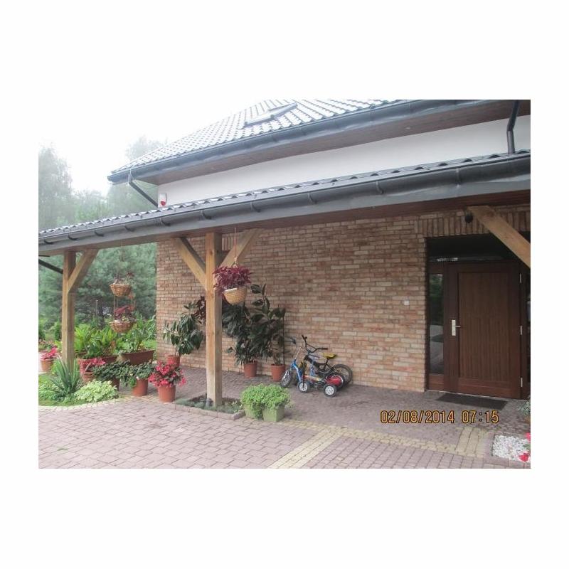 Dom jednorodzinny z ceglaną elewacją, drewnianą pergolą i roślinami w donicach, dwa dziecięce rowerki zaparkowane obok ściany.