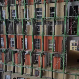 Budynek wielorodzinny w trakcie renowacji elewacji, osłonięty zielonym rusztowaniem, z widocznymi fragmentami nowej, pomarańczowej elewacji i elementami ocieplenia.