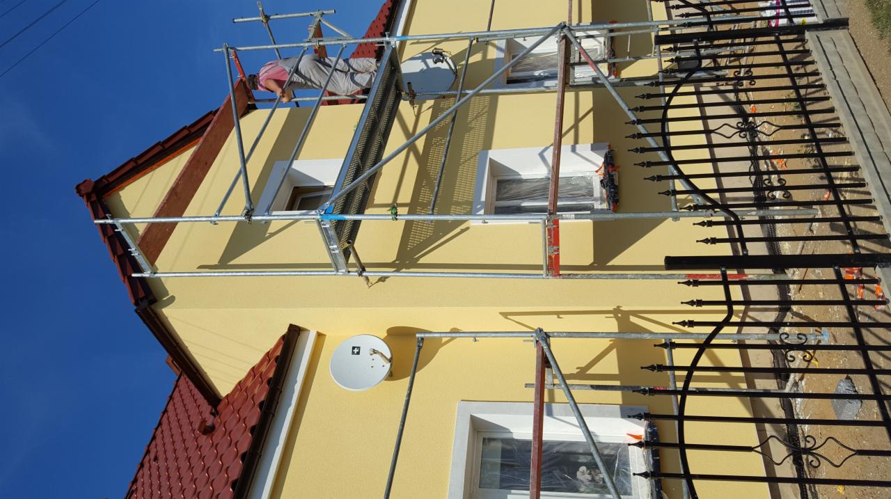 Pracownik na rusztowaniu maluje żółtą elewację domu jednorodzinnego, widoczna antena satelitarna i ogrodzenie z kutego żelaza.