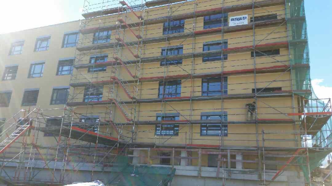 Budynek wielorodzinny w trakcie prac elewacyjnych, osłonięty rusztowaniami, widoczny pracownik na rusztowaniu. Na elewacji widoczny żółty tynk i logo firmy.