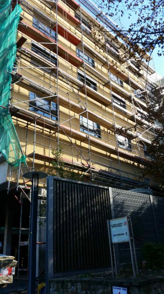 Budynek wielopiętrowy w trakcie prac elewacyjnych, otoczony rusztowaniami i zabezpieczony zieloną siatką ochronną, widoczny szyld informacyjny przed budynkiem.