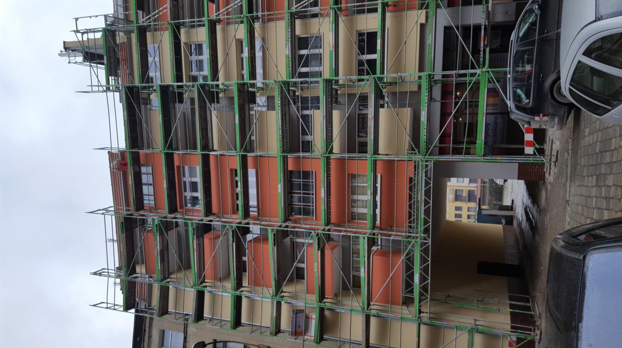 Budynek wielorodzinny w trakcie renowacji elewacji, osłonięty zielonym rusztowaniem, z widocznymi fragmentami nowej, pomarańczowej elewacji i elementami ocieplenia.