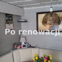 Jasnoszare wnętrze po renowacji z dekoracyjnym, kasetonowym sufitem, wiszącą lampą, drewnianą boazerią, narożną kanapą i okrągłym stołem z bukietem róż.