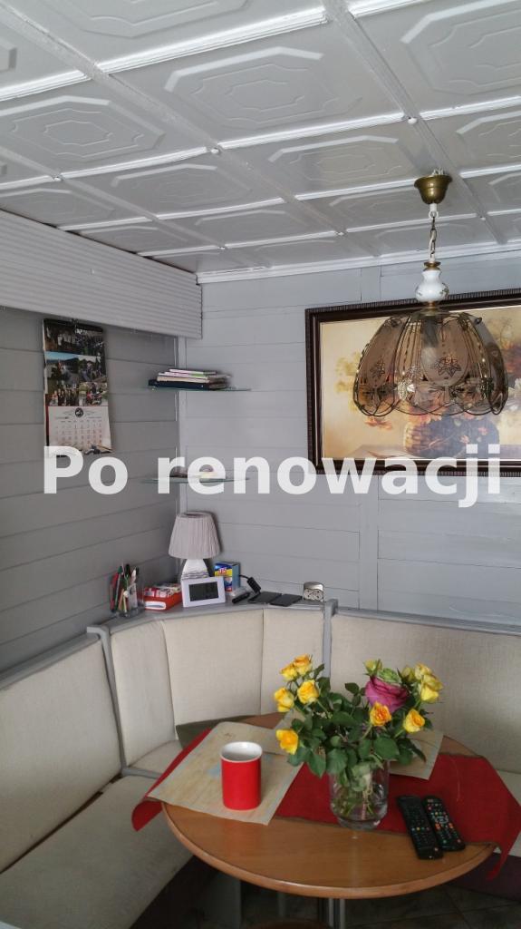 Jasnoszare wnętrze po renowacji z dekoracyjnym, kasetonowym sufitem, wiszącą lampą, drewnianą boazerią, narożną kanapą i okrągłym stołem z bukietem róż.