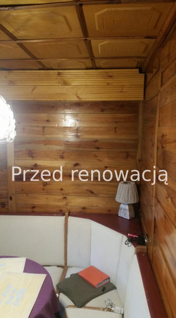 Pokój wyłożony boazerią przed renowacją, widoczny narożnik z sofą, lampka nocna i stół z fioletowym obrusem.