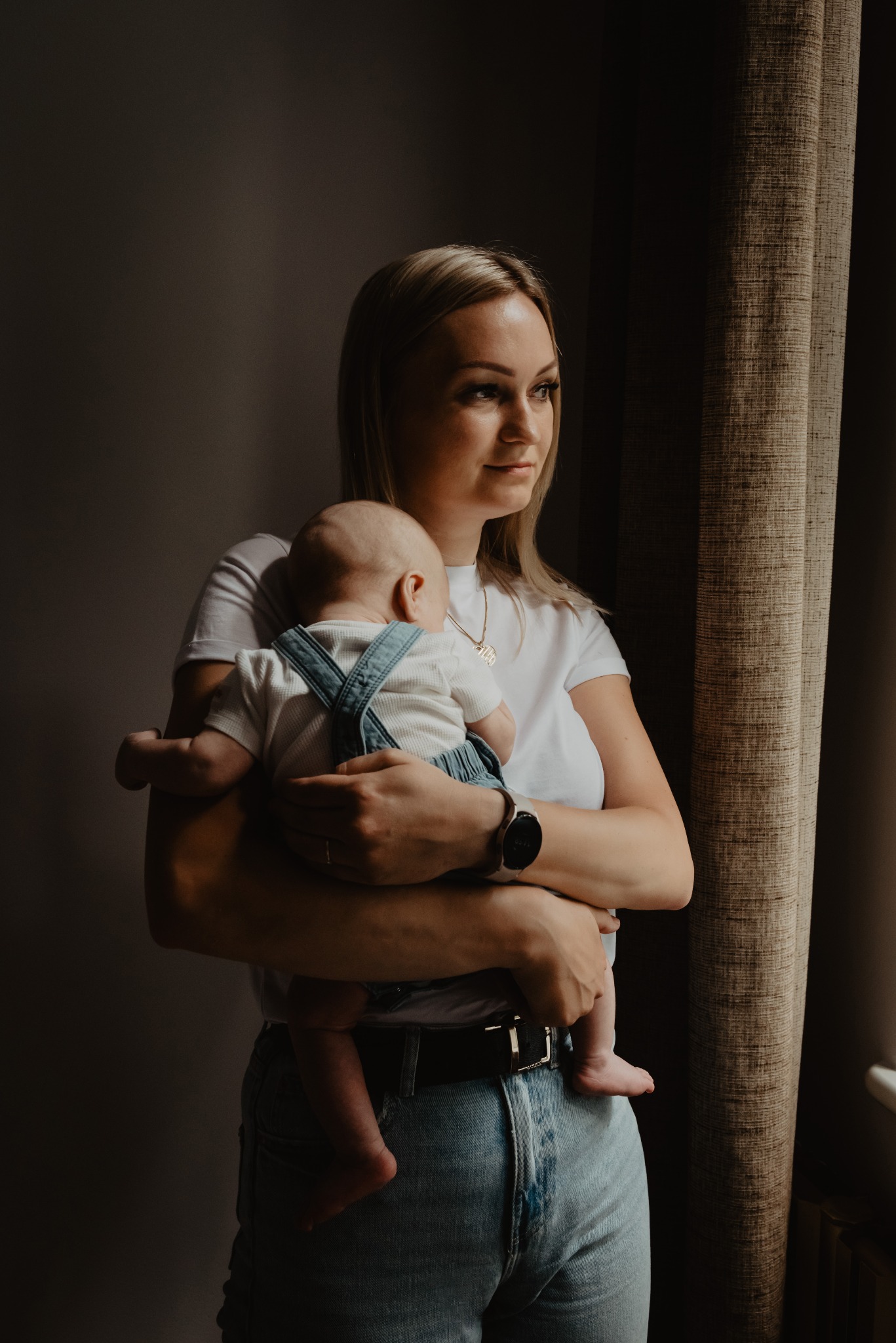 Kobieta z blond włosami w białej koszulce i jeansach trzymająca niemowlę w denimowych ogrodniczkach, oboje patrzą w stronę okna z miękkim światłem, obok teksturowana zasłona.