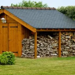 Drewniana drewutnia z szarym dachem krytym dachówką, z zapasem pociętego drewna opałowego, oświetlona lampą na ścianie frontowej, otoczona zielenią.