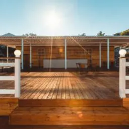 Drewniany taras z białymi balustradami prowadzący do zadaszonej konstrukcji z drewnianymi ścianami, oświetlony porannym słońcem.