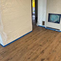 Lasmet - Pokój zabezpieczony folią i taśmą malarską przed malowaniem lub innymi pracami wykończeniowymi, widoczna drewnopodobna podłoga.