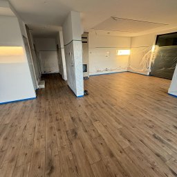 Lasmet - Przygotowane do malowania wnętrze z drewnopodobną podłogą, zabezpieczone folią i taśmą malarską, widoczny fragment sufitu podwieszanego.