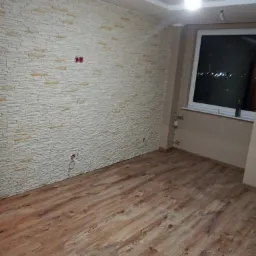 Pokój po remoncie z panelami podłogowymi w odcieniach brązu i ścianą wyłożoną dekoracyjną okładziną imitującą jasny kamień, widoczne gniazdka elektryczne i okno z widokiem na nocne miasto.