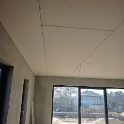 Montaż płyt gipsowo-kartonowych na suficie w surowym wnętrzu, widoczne łączenia płyt i wkręty, przez okno widok na budynek w trakcie budowy.