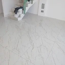 Nowo położona, błyszcząca podłoga z jasnych płytek imitujących marmur, widok na surowe schody z zieloną barierką, na których leżą worki z zaprawą i ubrania.