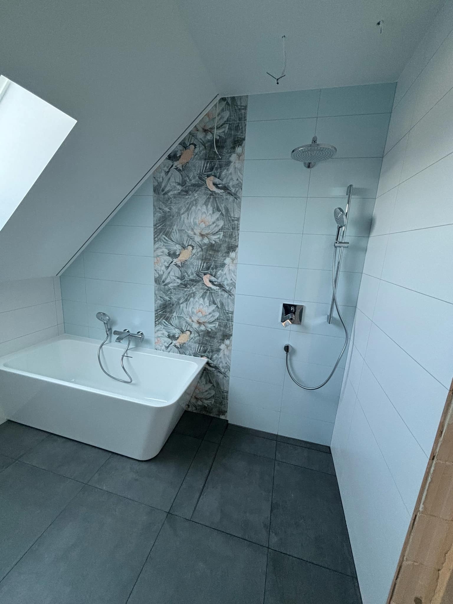 Nowoczesna łazienka na poddaszu z białą wanną, szarą podłogą z płytek i akcentem dekoracyjnym w postaci pionowego pasa płytek z motywem ptaków i roślin, obok prysznic z deszczownicą.