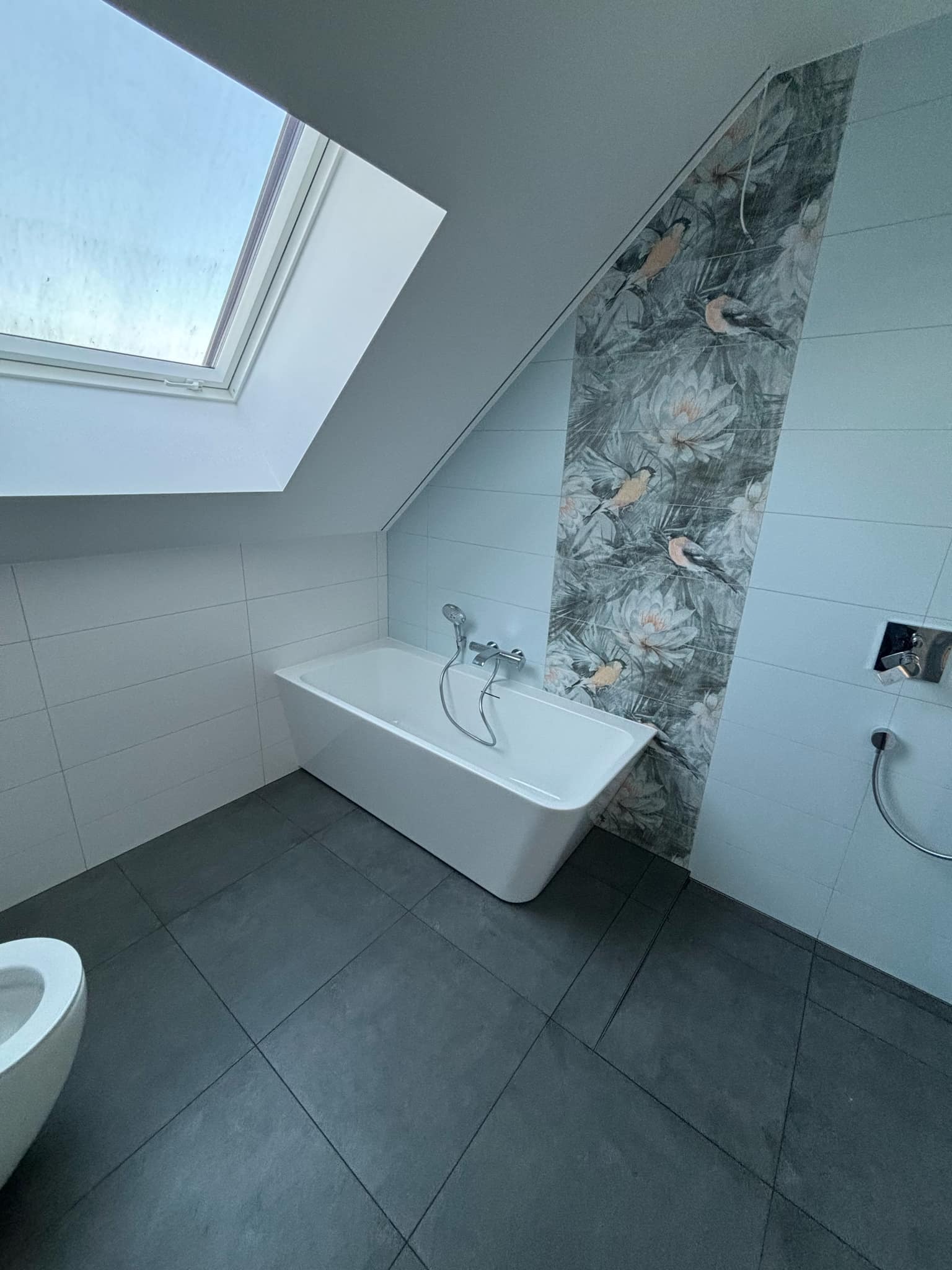 Nowoczesna łazienka na poddaszu z białą wanną wolnostojącą, szarą podłogą z płytek, białą glazurą i dekoracyjnym pasem płytek z motywem ptaków i kwiatów, oświetlona oknem dachowym.