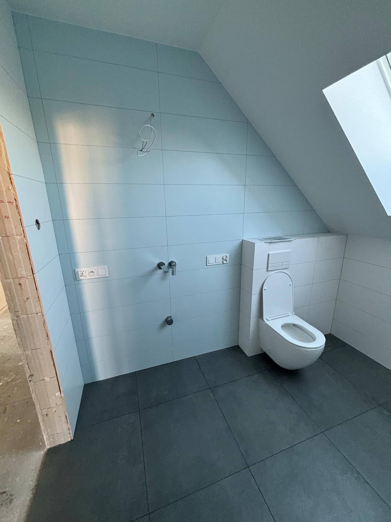 Ukończona łazienka na poddaszu ze skośnym sufitem, wyłożona jasnoniebieskimi płytkami ściennymi i ciemnoszarą podłogą, z zamontowaną białą toaletą i widocznymi instalacjami.