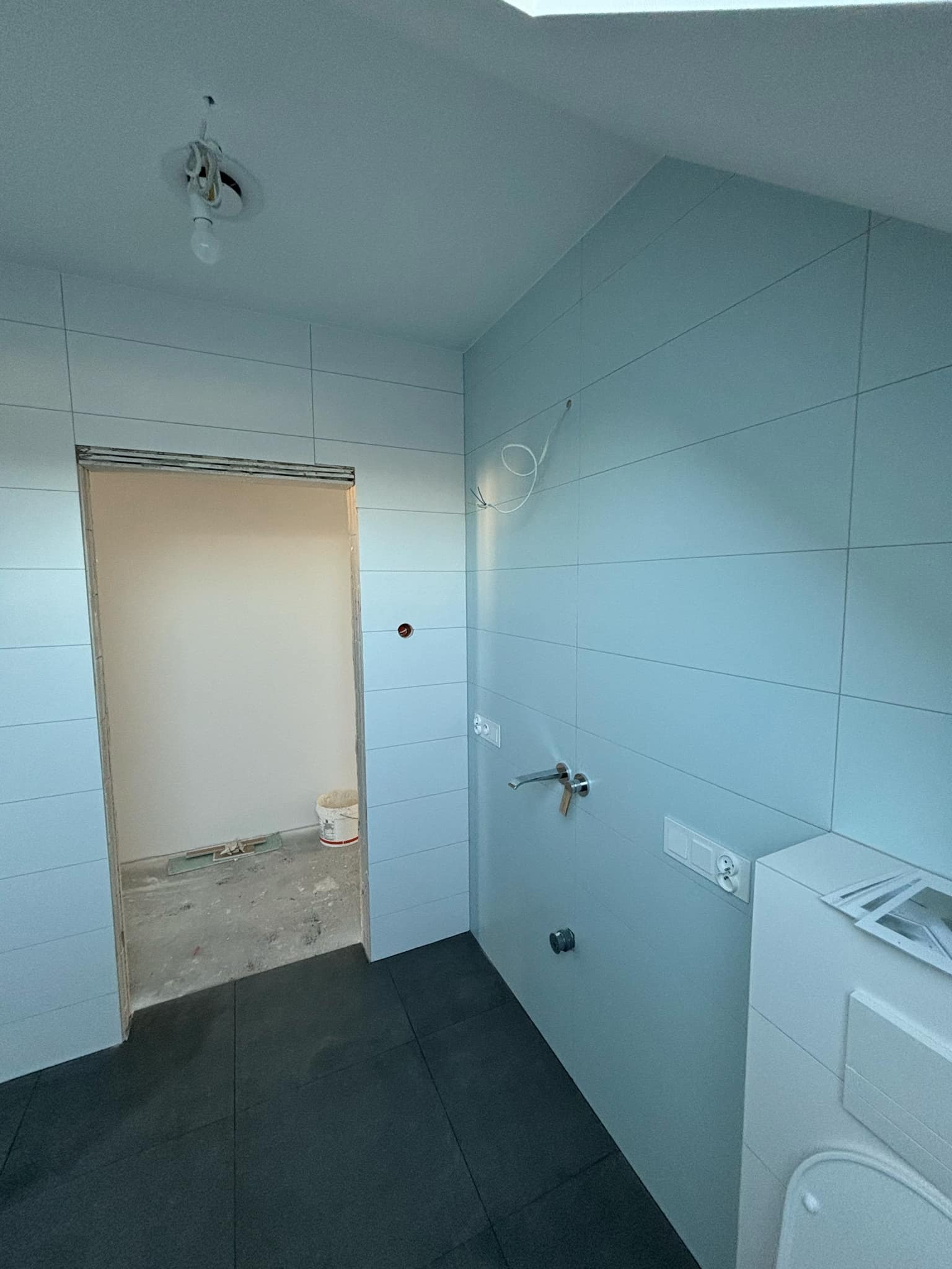 Łazienka w trakcie remontu: jasnoniebieskie płytki ścienne, czarne płytki podłogowe, surowa instalacja elektryczna, widoczny fragment armatury i otwór drzwiowy do pomieszczenia z wiadrem...