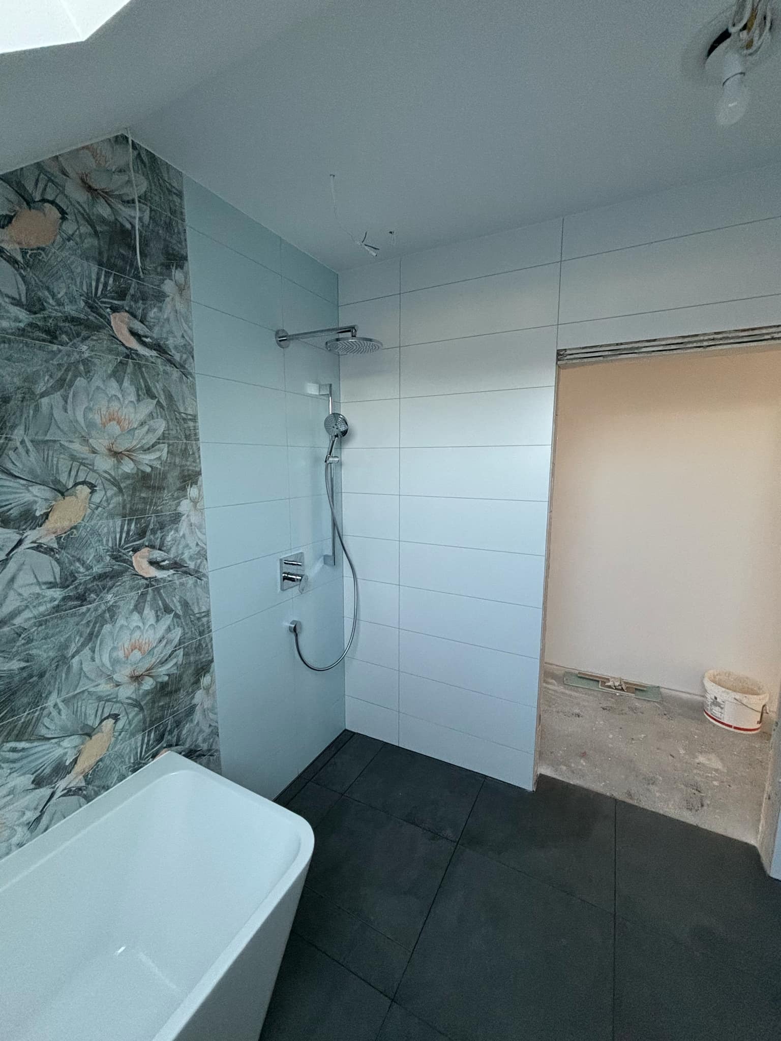 Nowoczesna łazienka w trakcie wykańczania: biała wanna, płytki z motywem ptaków, szare płytki podłogowe, białe płytki ścienne i chromowana armatura prysznicowa. Widoczny fragment sufitu...