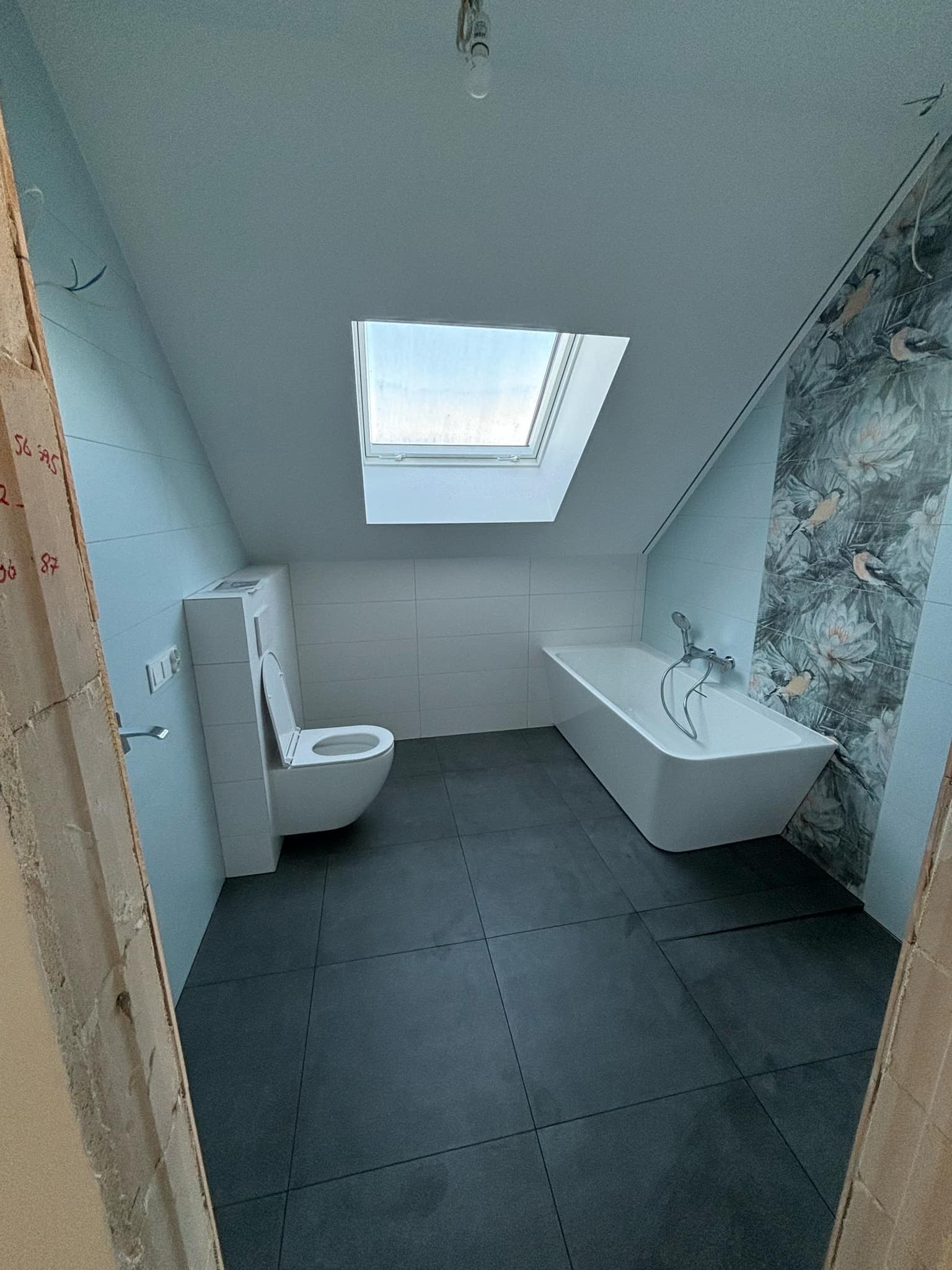 Ukończona łazienka na poddaszu ze skośnym dachem, z oknem dachowym, białą wanną i toaletą w zabudowie, ciemną podłogą z płytek i ścianą z motywem kwiatowo-ptasim.