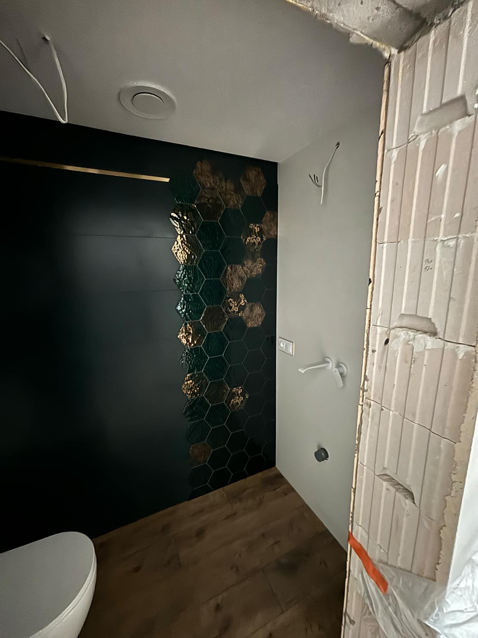 Wnętrze łazienki w trakcie remontu: widoczna ściana z ciemnymi, heksagonalnymi płytkami z akcentami złota, surowa ściana z cegieł, drewnopodobna podłoga i fragment białej umywalki.