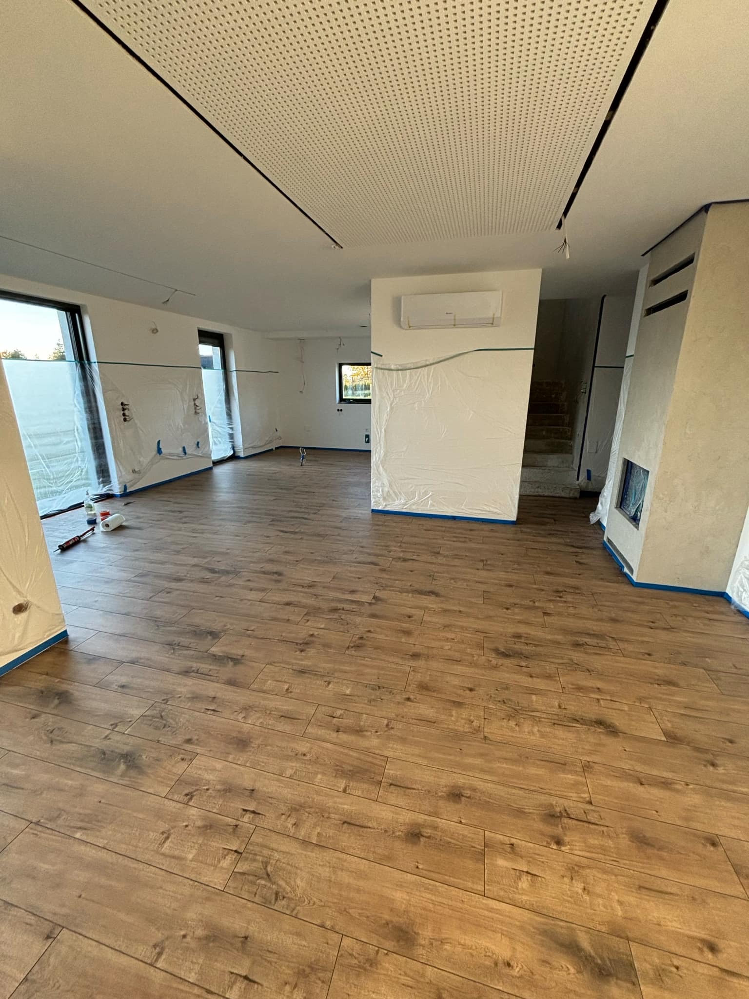 Pokój w trakcie przygotowań do malowania: zabezpieczone folią okna, ściany i klimatyzator, drewnopodobna podłoga, perforowany sufit podwieszany.
