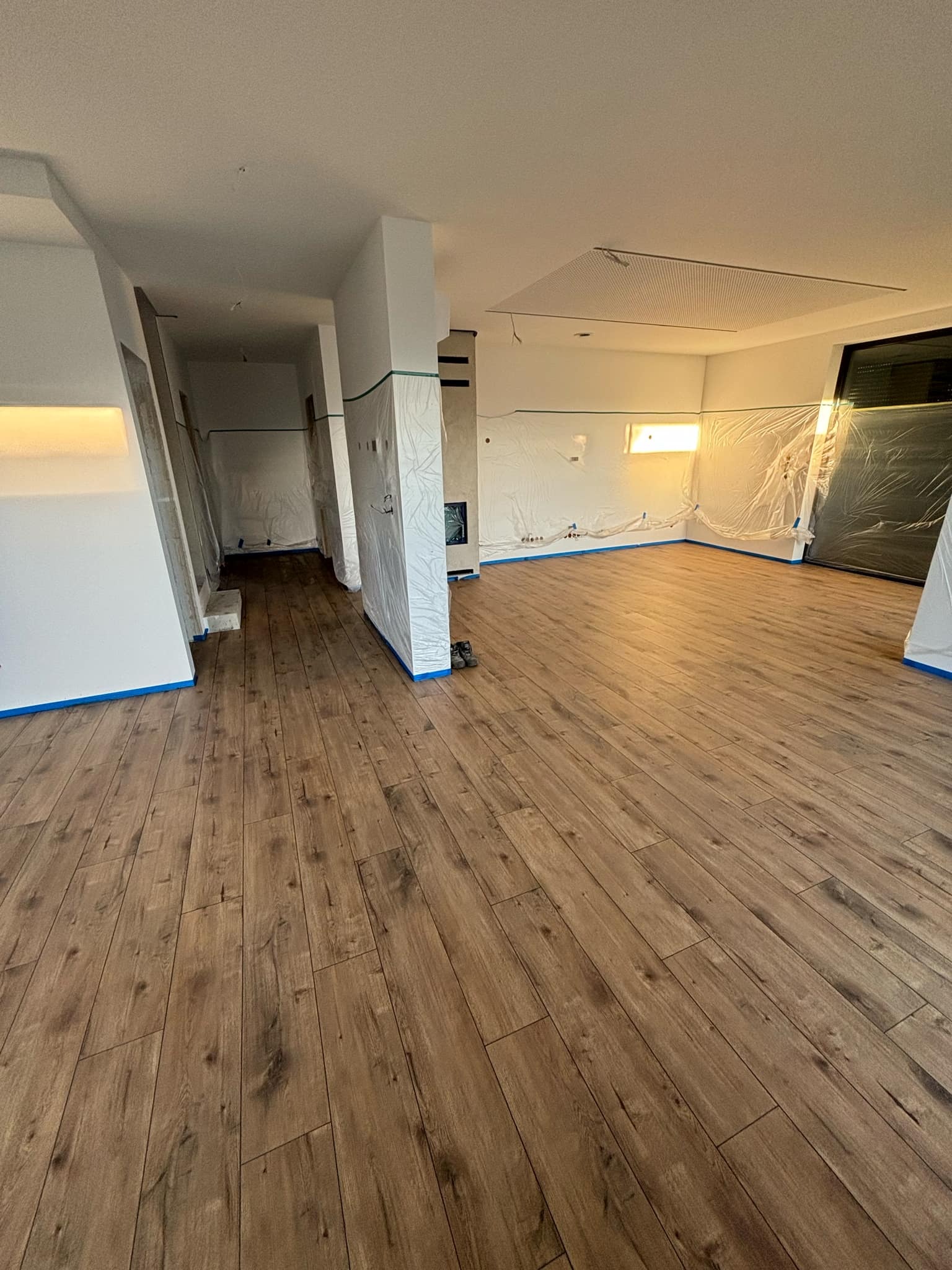 Przygotowane do malowania wnętrze z drewnopodobną podłogą, zabezpieczone folią i taśmą malarską, widoczny fragment sufitu podwieszanego.