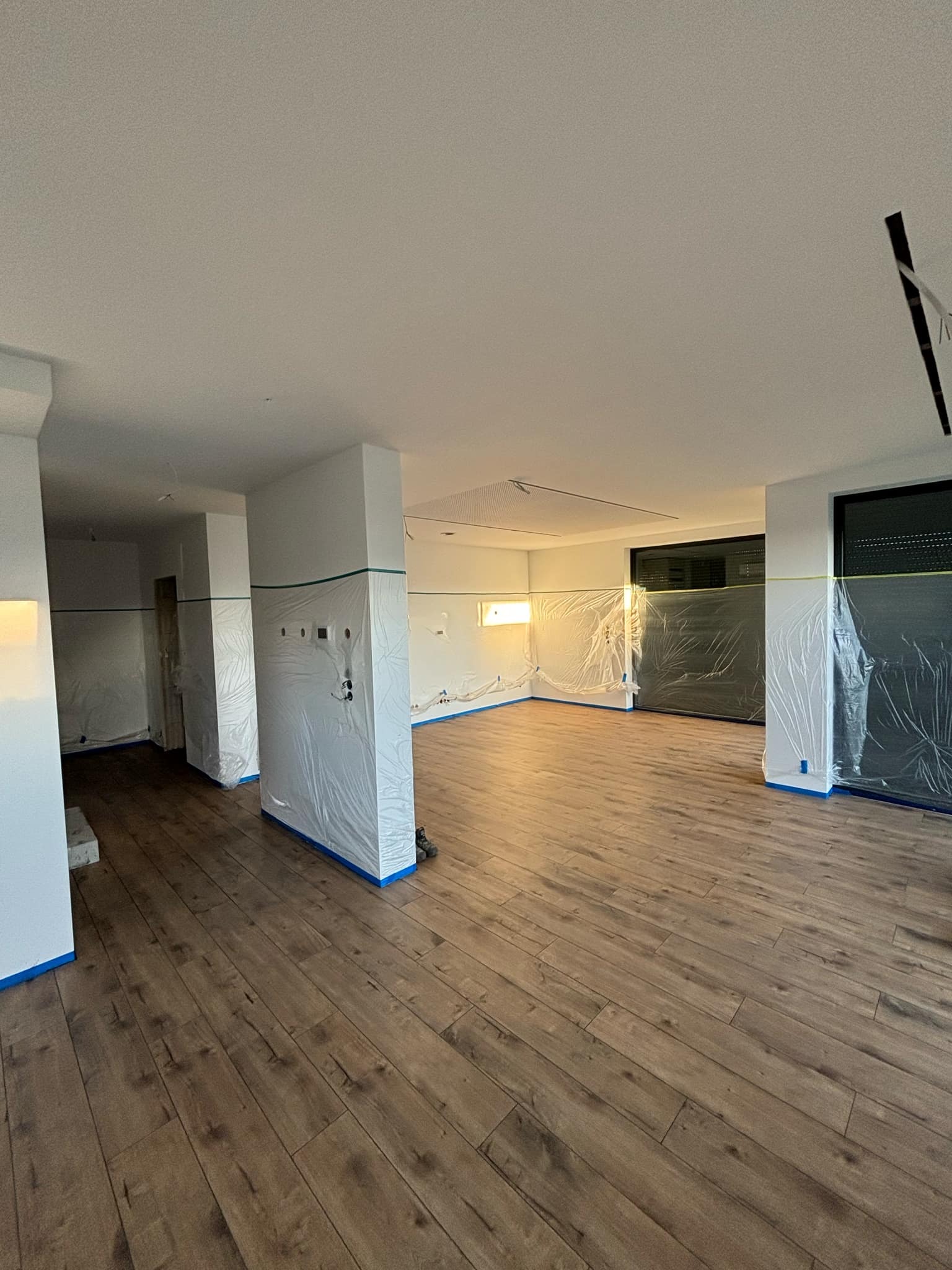 Przygotowane do malowania wnętrze z drewnianą podłogą, ściany i okna zabezpieczone folią i niebieską taśmą malarską.