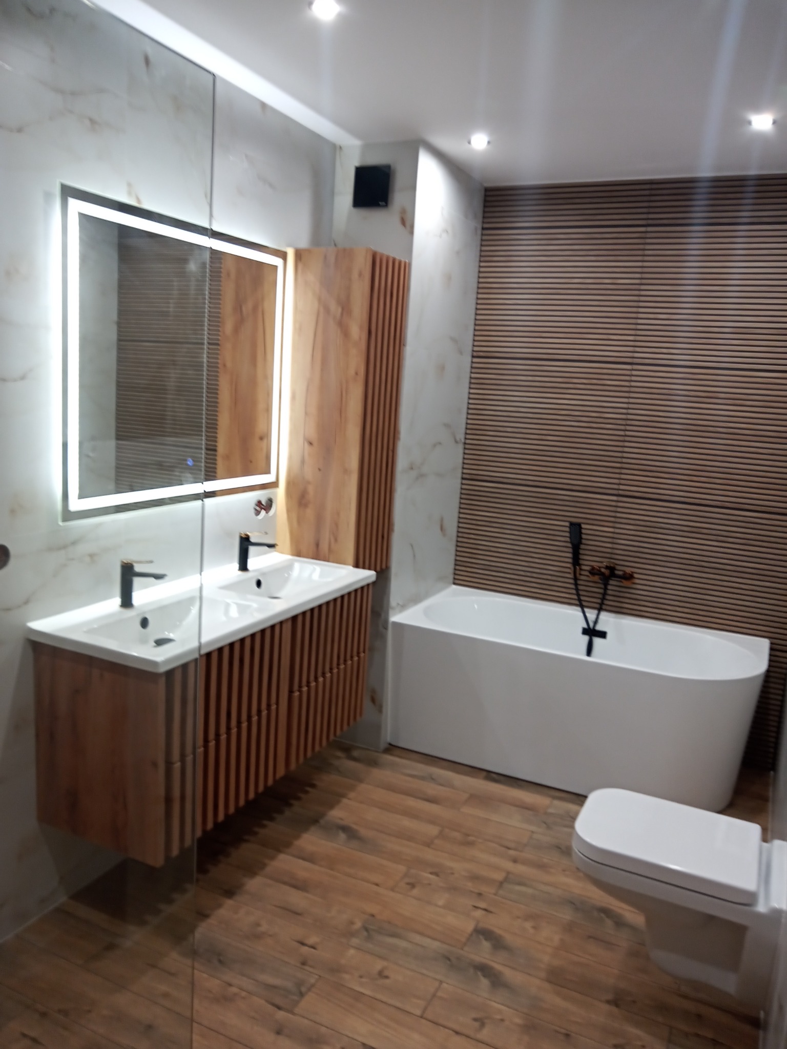 Nowoczesna łazienka z drewnianymi akcentami: szafka pod umywalkę z lamelami, podświetlane lustro, wolnostojąca wanna i poziome panele ścienne.