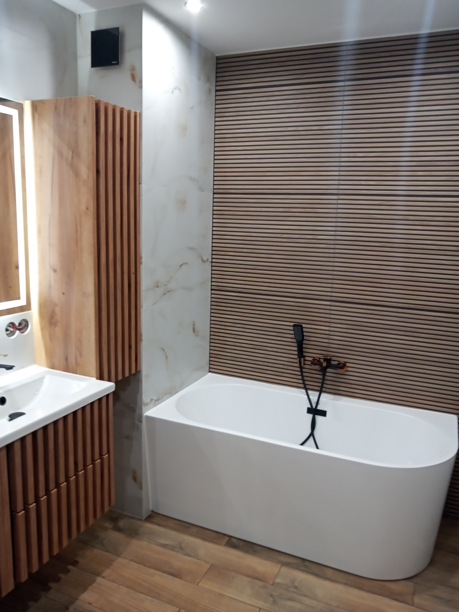 Nowoczesna łazienka z drewnianymi lamelami na ścianie, białą wanną wolnostojącą z czarną armaturą, umywalką z szafką w stylu lamelowym i płytkami imitującymi marmur.