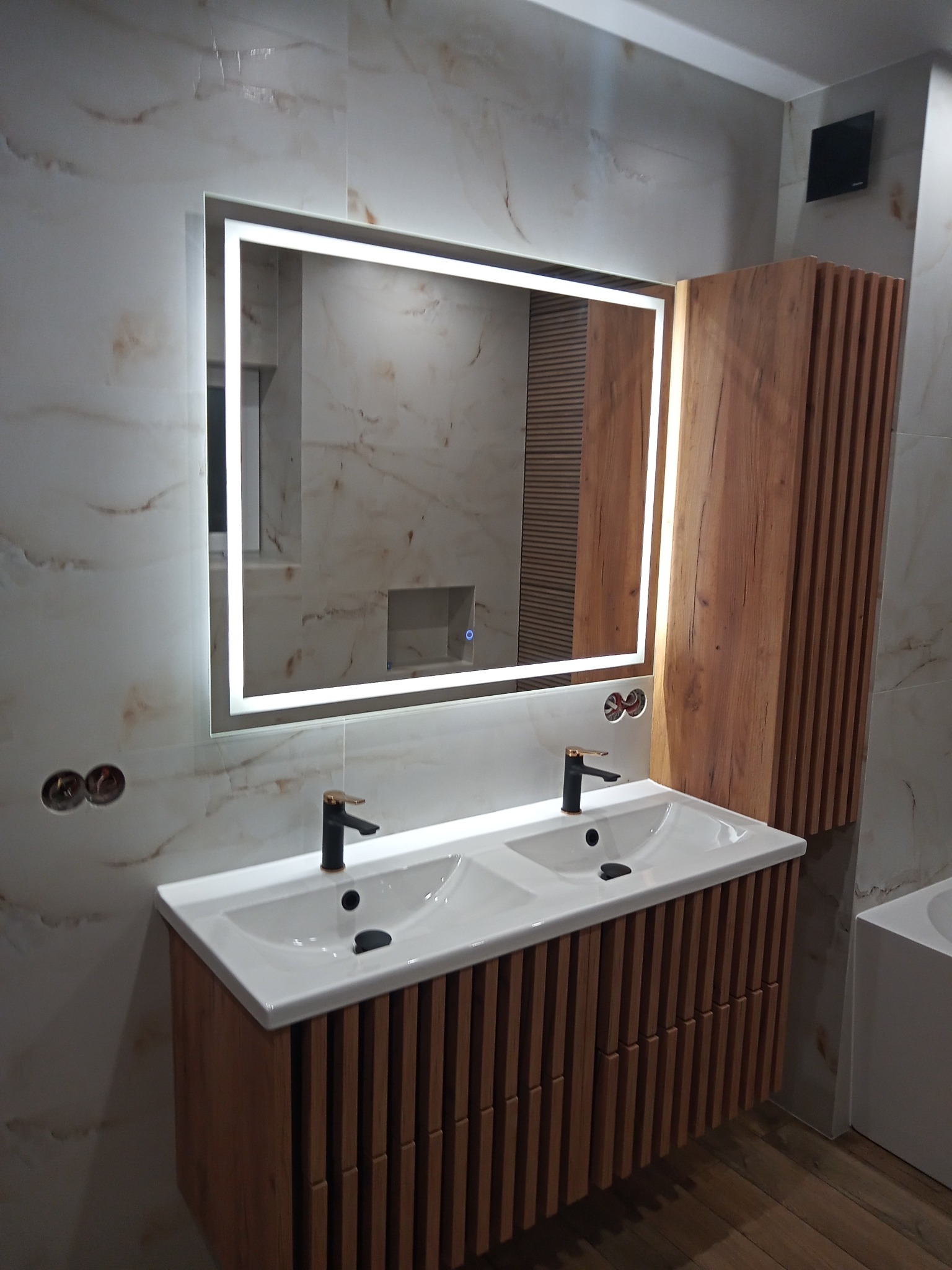 Nowoczesna łazienka z podświetlanym lustrem, umywalką nablatową z dwiema misami i drewnianą szafką z lamelami, marmurowe płytki na ścianach.