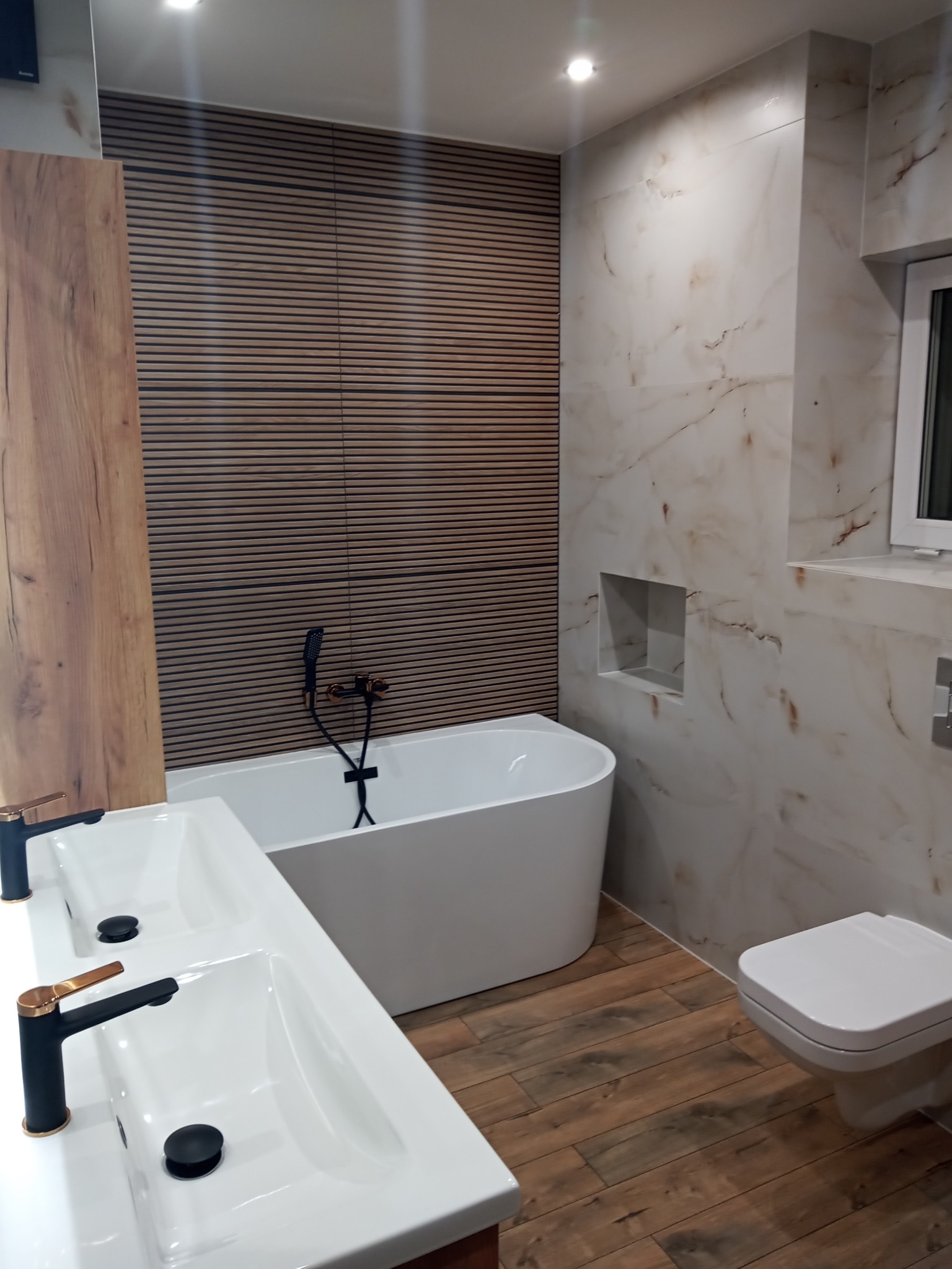 Nowoczesna łazienka z wolnostojącą wanną, podwójną umywalką z czarno-złotą armaturą, drewnianymi panelami ściennymi i marmurowymi płytkami.