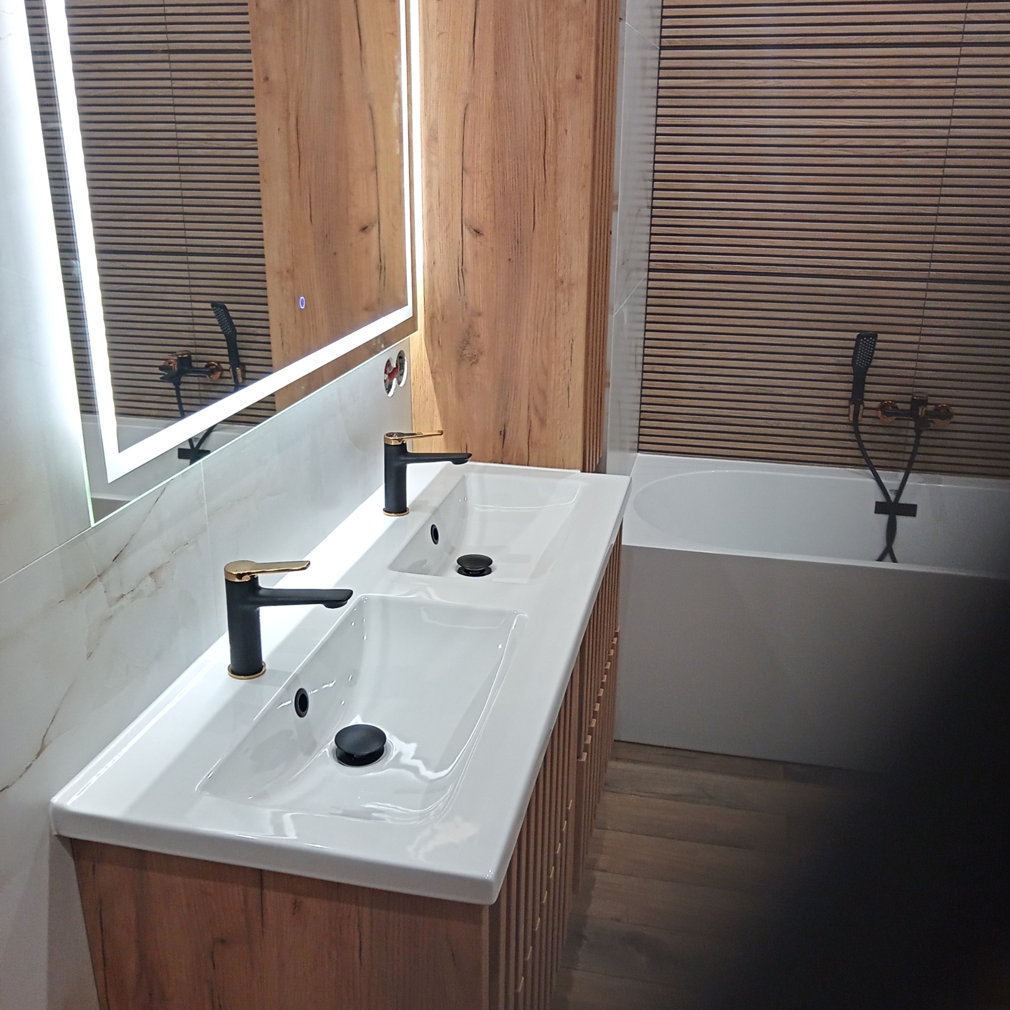 Nowoczesna łazienka w Mściszewicach z podwójną umywalką na drewnianej szafce, armatura w kolorze czarnym ze złotymi akcentami, lustro z podświetleniem LED i drewniana okładzina ścienna.