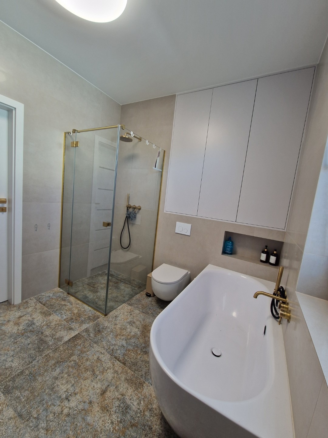 Nowoczesna łazienka z kabiną prysznicową w złotej ramie, wolnostojącą białą wanną i beżowymi płytkami podłogowymi o wzorzystej fakturze.