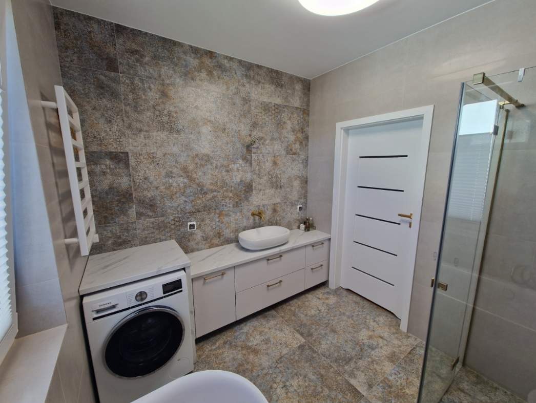 Nowoczesna łazienka z płytkami imitującymi kamień, białą armaturą, pralką zabudowaną pod blatem i kabiną prysznicową ze szklanymi drzwiami.