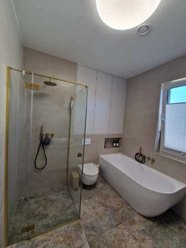 Nowoczesna łazienka z kabiną prysznicową typu walk-in z złotymi profilami, wolnostojącą wanną i płytkami imitującymi kamień na podłodze.