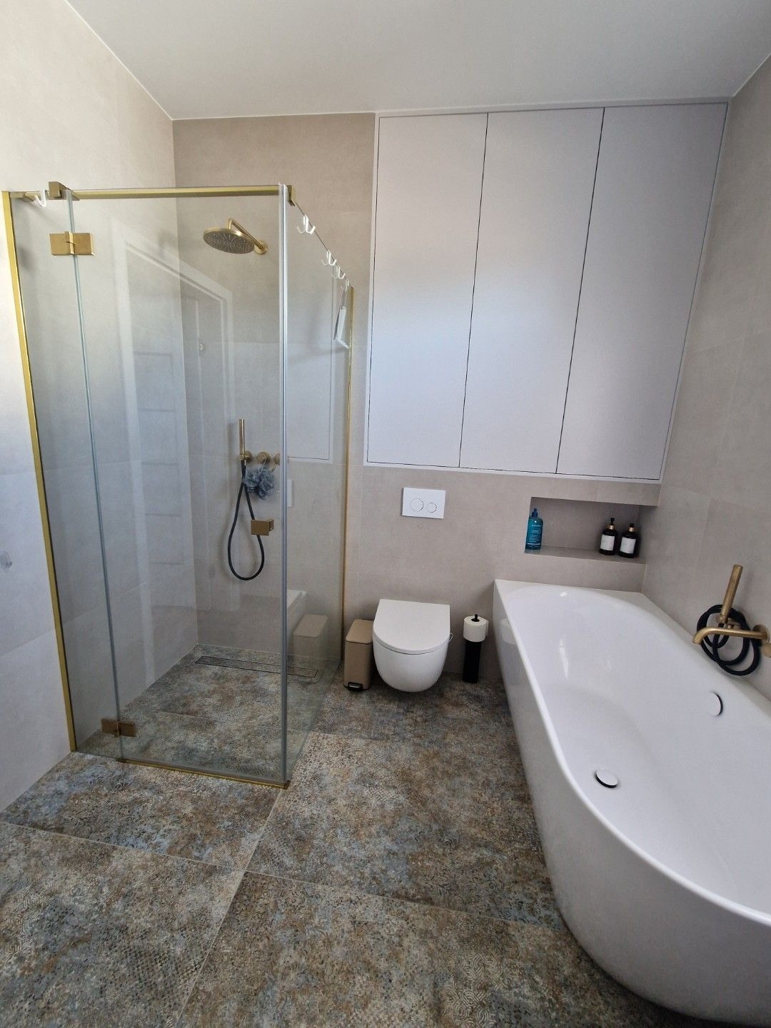 Kompleksowo wykończona łazienka w beżowej kolorystyce z kabiną prysznicową ze złotymi detalami, białą wanną, podwieszaną toaletą i wzorzystą podłogą z płytek.