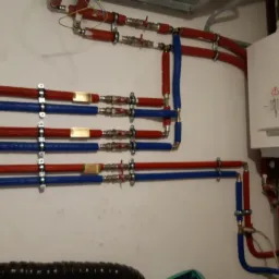 Instalacja grzewcza z kotłem kondensacyjnym, widoczne rury w kolorze czerwonym i niebieskim, połączone z zaworami i przymocowane do ściany za pomocą metalowych uchwytów.