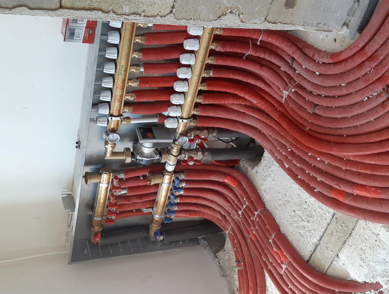 Rozdzielacz ogrzewania podłogowego z czerwonymi rurami w izolacji, widoczne zawory i manometry, montaż w surowym stanie.