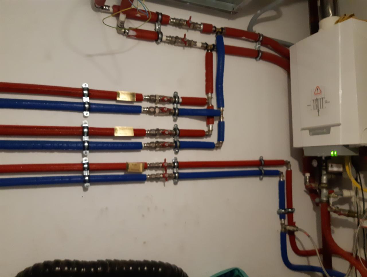 Instalacja grzewcza z kotłem kondensacyjnym, widoczne rury w kolorze czerwonym i niebieskim, połączone z zaworami i przymocowane do ściany za pomocą metalowych uchwytów.
