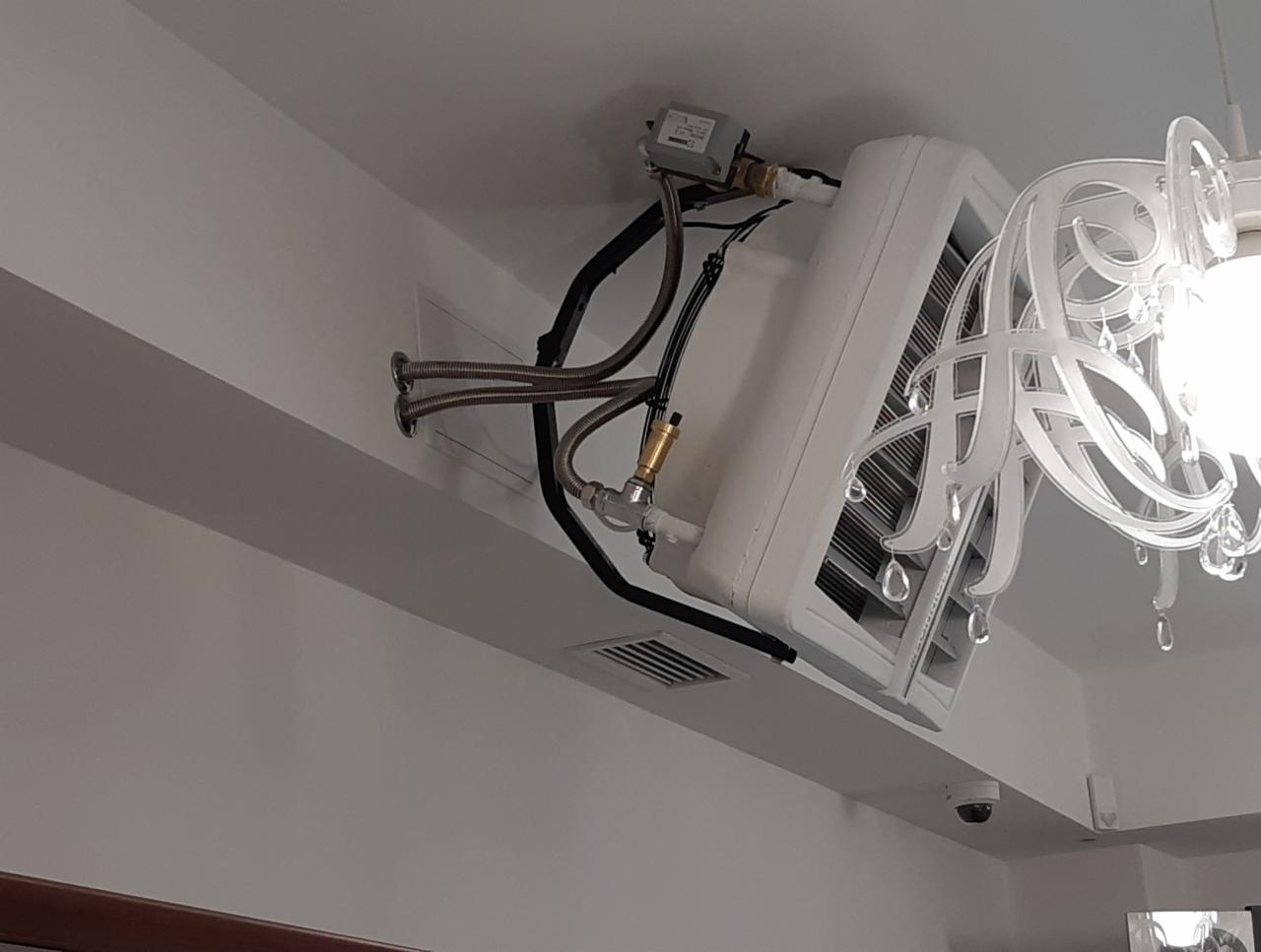 Biały klimakonwektor kanałowy zamontowany pod sufitem, widoczne podłączenia hydrauliczne i elektryczne, obok ozdobny żyrandol.